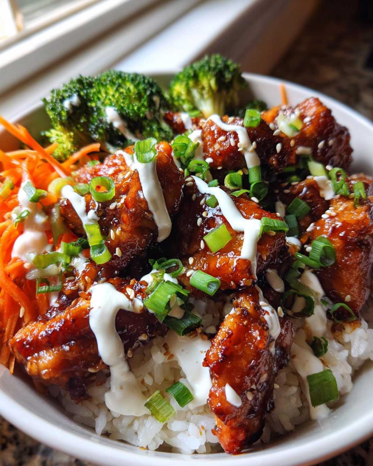 Close-up of a Honey Soy Chicken Bowl featuring glazed chicken pieces over rice, topped with white sauce, scallions, and sesame seeds.