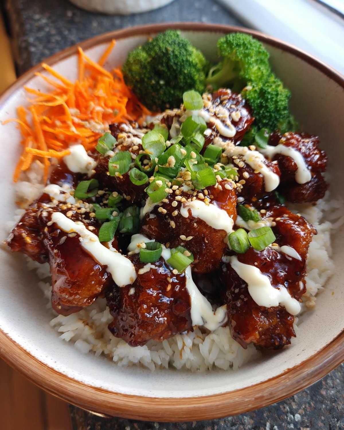 Close-up of a Honey Soy Chicken Bowl featuring glazed chicken pieces over rice, topped with white sauce, scallions, and sesame seeds.