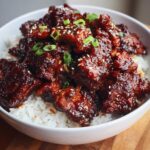 Close-up of a white bowl filled with white rice topped generously with glistening, dark Sticky BBQ Chicken Bowl with Rice, garnished with sesame seeds and green onions.