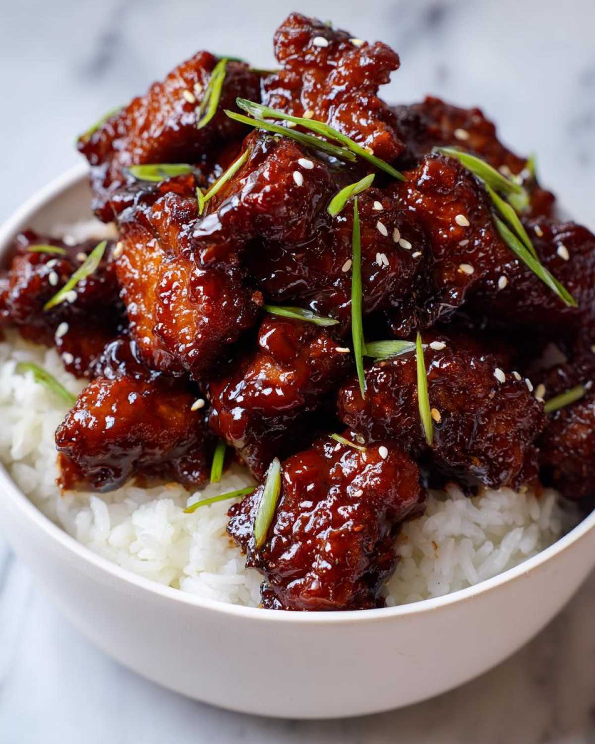 Close-up of a white bowl filled with white rice topped generously with sticky BBQ chicken pieces and garnished with sesame seeds and green onions.