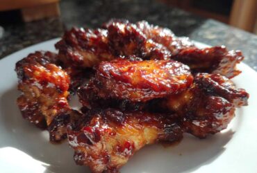A close-up of several Sticky BBQ Chicken Wings Crispy Oven Style coated in a thick, glossy sauce on a white plate.