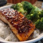 A perfectly cooked fillet of Sticky Honey Soy Salmon glazed with sauce and sesame seeds, served over white rice with steamed broccoli.