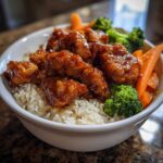 A close-up of a white bowl filled with white rice topped with glossy Sticky Orange Chicken Bowl with Rice and Veggies and steamed broccoli and carrots.