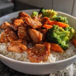 A close-up of a white bowl filled with Sticky Orange Chicken Bowl with Rice and Veggies, featuring glazed chicken pieces over white rice and bright green broccoli florets.