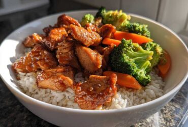 A close-up of a white bowl filled with Sticky Orange Chicken Bowl with Rice and Veggies, featuring glazed chicken pieces over white rice and bright green broccoli florets.