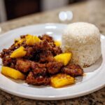 Close-up of Sticky Pineapple Chicken with a sweet glaze served next to a molded serving of white rice on a white plate.