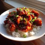 A close-up of Sticky Sweet Chili Chicken Skillet pieces coated in glossy sauce, served over white rice and garnished with sesame seeds and green onions.