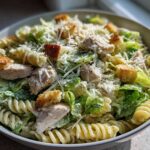 A bowl filled with Chicken Caesar Pasta Salad featuring rotini pasta, grilled chicken pieces, romaine lettuce, croutons, and grated Parmesan cheese.