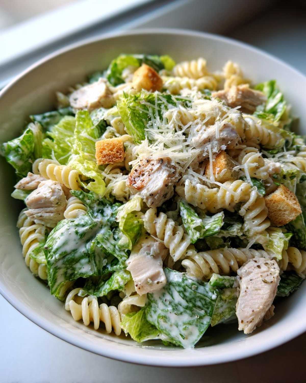 A close-up overhead view of a bowl filled with Chicken Caesar Pasta Salad, featuring rotini pasta, romaine lettuce, chicken pieces, croutons, and grated cheese.