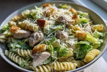 A bowl filled with Chicken Caesar Pasta Salad featuring rotini pasta, grilled chicken pieces, romaine lettuce, croutons, and grated Parmesan cheese.