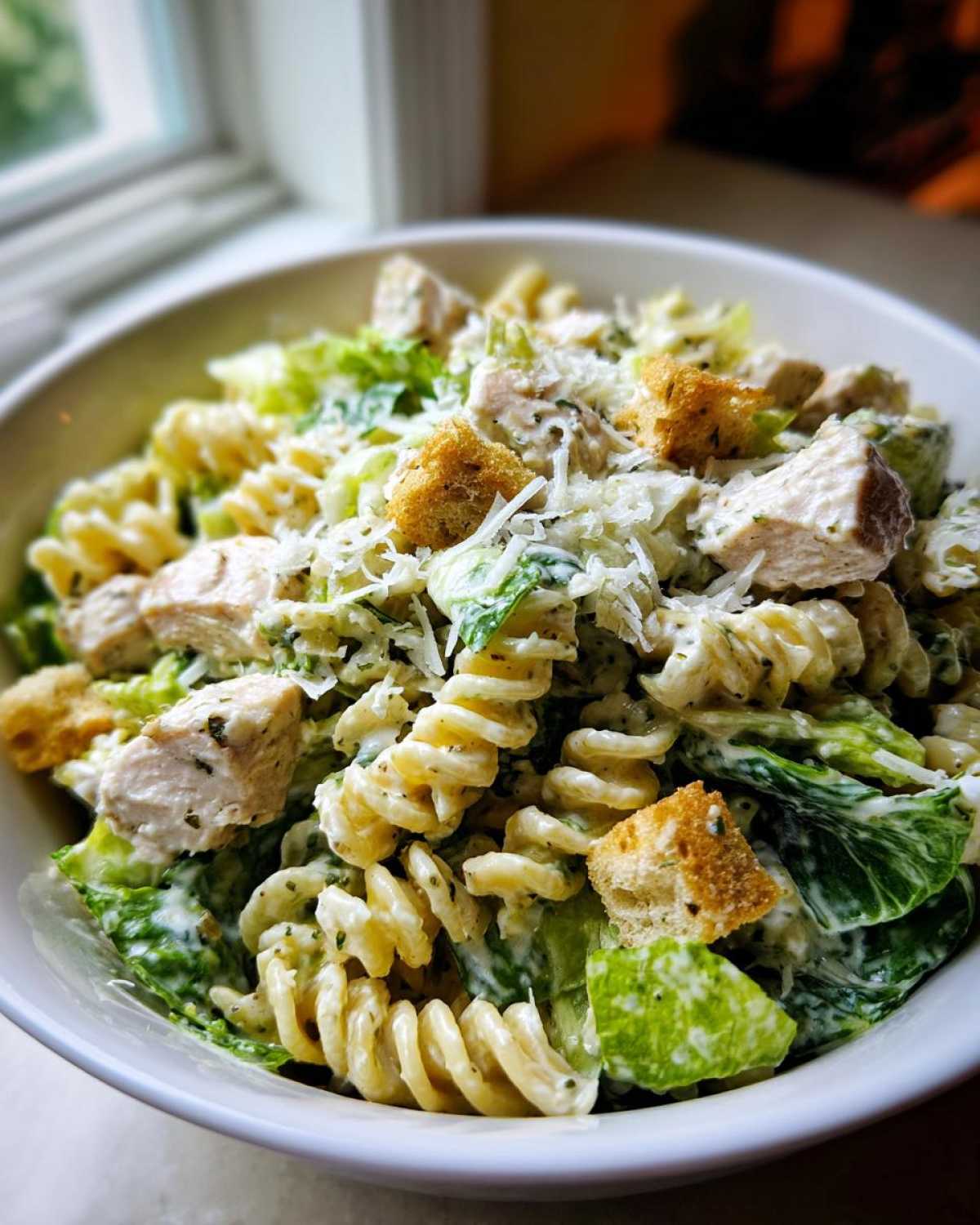 A close-up of a bowl filled with creamy Chicken Caesar Pasta Salad featuring rotini pasta, chicken chunks, romaine lettuce, croutons, and grated Parmesan.