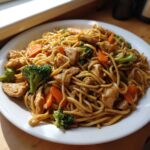 A plate piled high with savory Stir-Fried Noodles featuring chicken pieces, bright green broccoli florets, and sliced carrots.