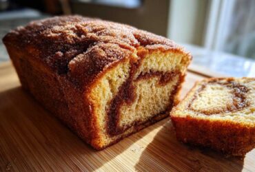 A loaf of Cinnamon Sugar Donut Bread, partially sliced, resting on a wooden cutting board in sunlight.
