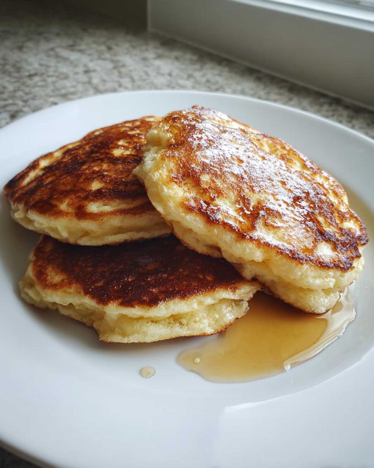 Three golden-brown Cottage Cheese Pancakes stacked on a white plate with a drizzle of maple syrup.