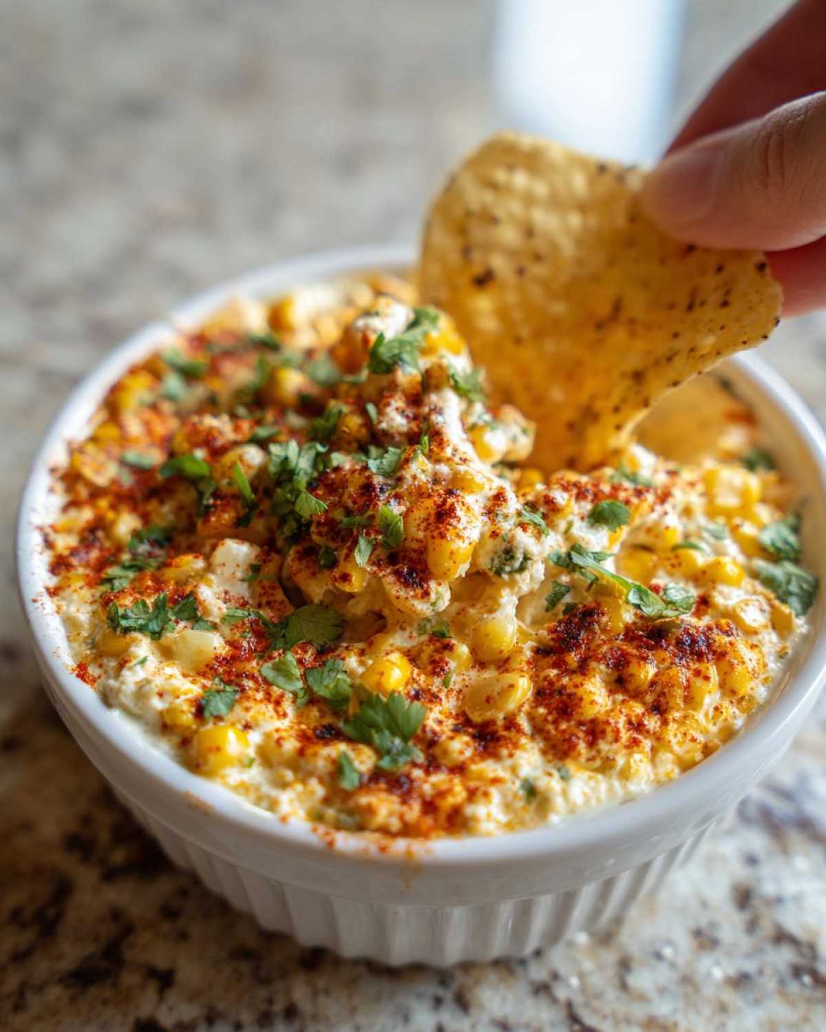 A hand dips a tortilla chip into a white bowl filled with creamy Elote Dip, topped with chili powder and cilantro.