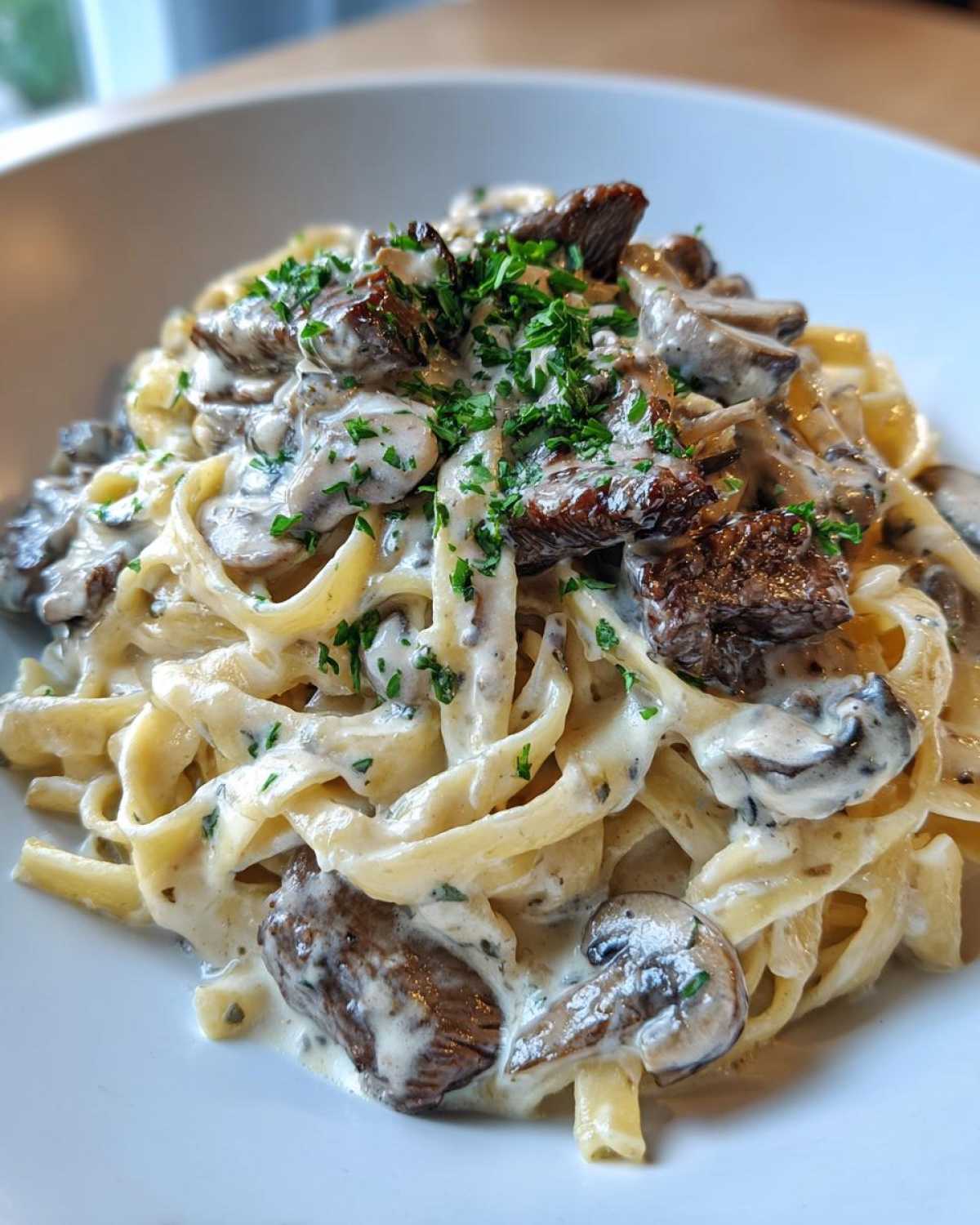 Close-up of creamy steak pasta featuring fettuccine, rich garlic cream sauce, steak pieces, and mushrooms, garnished with parsley.