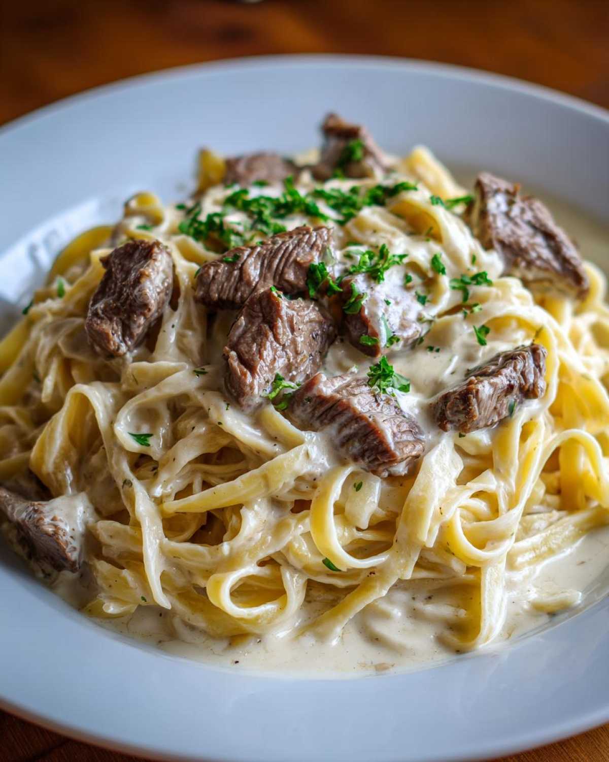 Close-up of a bowl of Creamy Steak Pasta featuring fettuccine noodles coated in a rich white sauce and topped with chunks of steak.