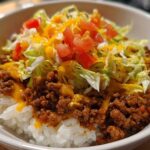 Close-up of a white bowl filled with Taco Rice: seasoned ground beef over white rice, topped with shredded lettuce, diced tomatoes, and melted cheese.