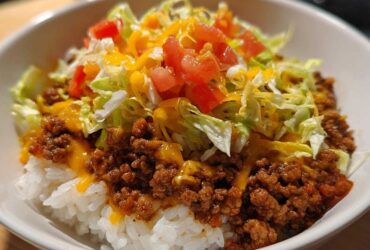 Close-up of a white bowl filled with Taco Rice: seasoned ground beef over white rice, topped with shredded lettuce, diced tomatoes, and melted cheese.