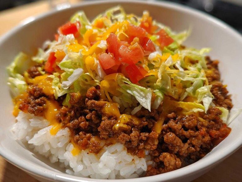 Close-up of a white bowl filled with Taco Rice: seasoned ground beef over white rice, topped with shredded lettuce, diced tomatoes, and melted cheese.
