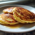Three golden-brown Cottage Cheese Pancakes stacked on a white plate, drizzled with maple syrup.
