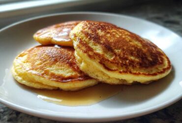 Three golden-brown Cottage Cheese Pancakes stacked on a white plate, drizzled with maple syrup.