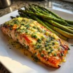 Close-up of a perfectly cooked Garlic Butter Baked Salmon Dinner topped with melted butter, minced garlic, and parsley, served alongside roasted asparagus.