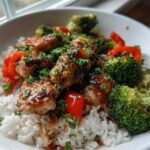A close-up of a white bowl filled with Garlic Chicken Bowls, featuring glazed chicken pieces over white rice with broccoli and red peppers.