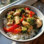 A close-up of a bowl filled with white rice, topped with seasoned chicken pieces, bright red bell peppers, and green broccoli florets, garnished with parsley, representing Garlic Chicken Bowls.