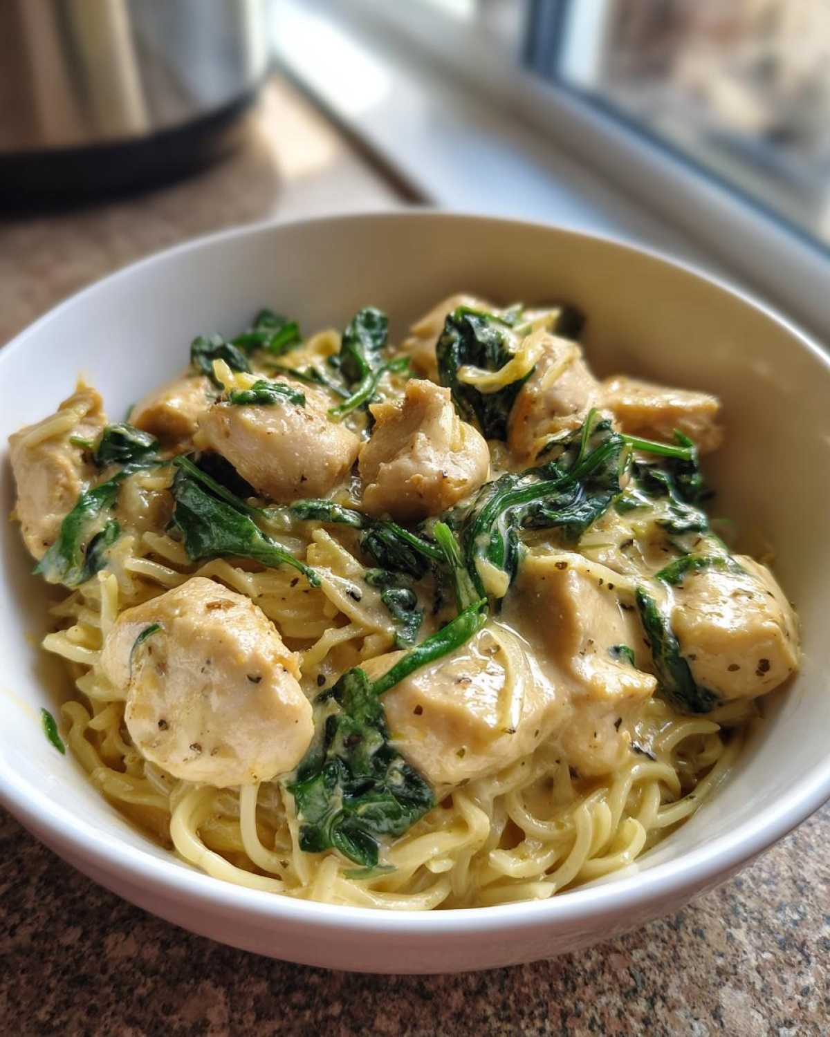A white bowl filled with Garlic Parmesan Chicken Ramen noodles, topped with creamy sauce, seasoned chicken pieces, and wilted spinach.
