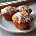 Three golden brown Apple Fritter Muffins drizzled with thick white glaze, sitting on a white plate near a window.