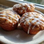 Three freshly baked Apple Fritter Muffins drizzled with white glaze, sitting on a speckled ceramic plate near a window.