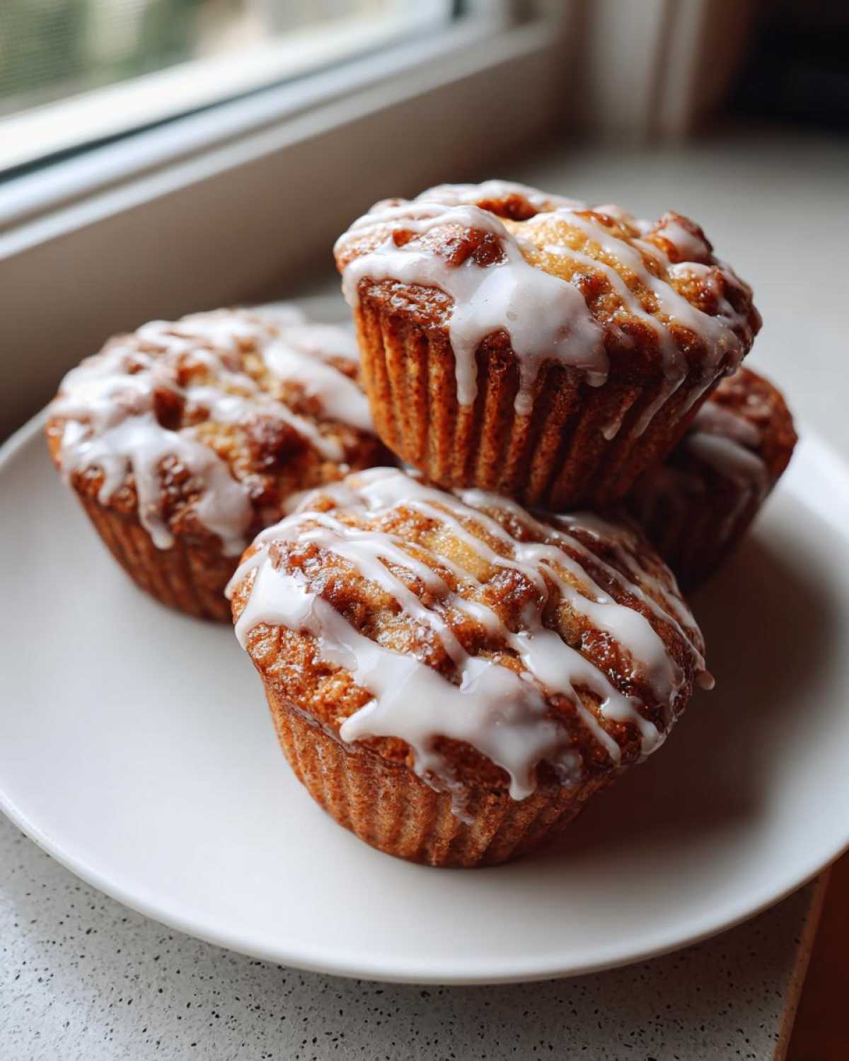 A stack of four freshly baked Apple Fritter Muffins drizzled with white glaze, resting on a white plate near a window.