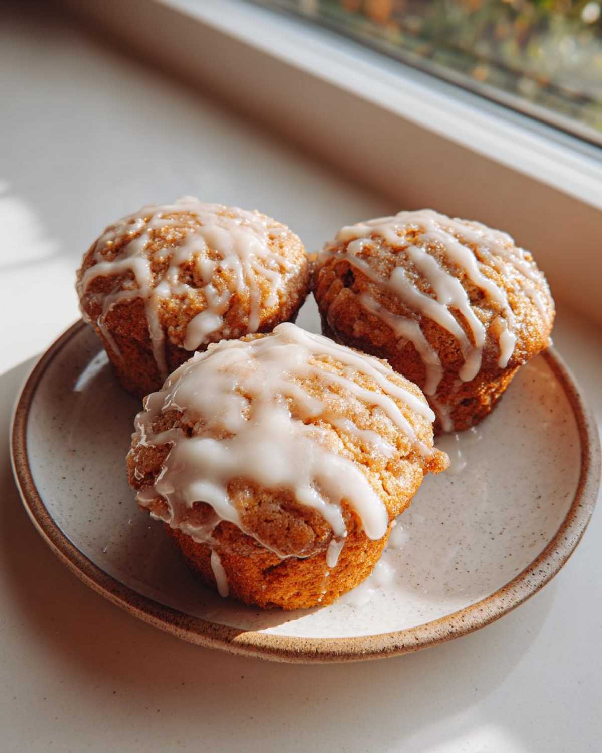 Three freshly baked Apple Fritter Muffins topped with a thick white glaze, sitting on a speckled ceramic plate near a window.