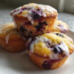 A stack of four golden Lemon Blueberry Muffins topped with a thick white lemon glaze.
