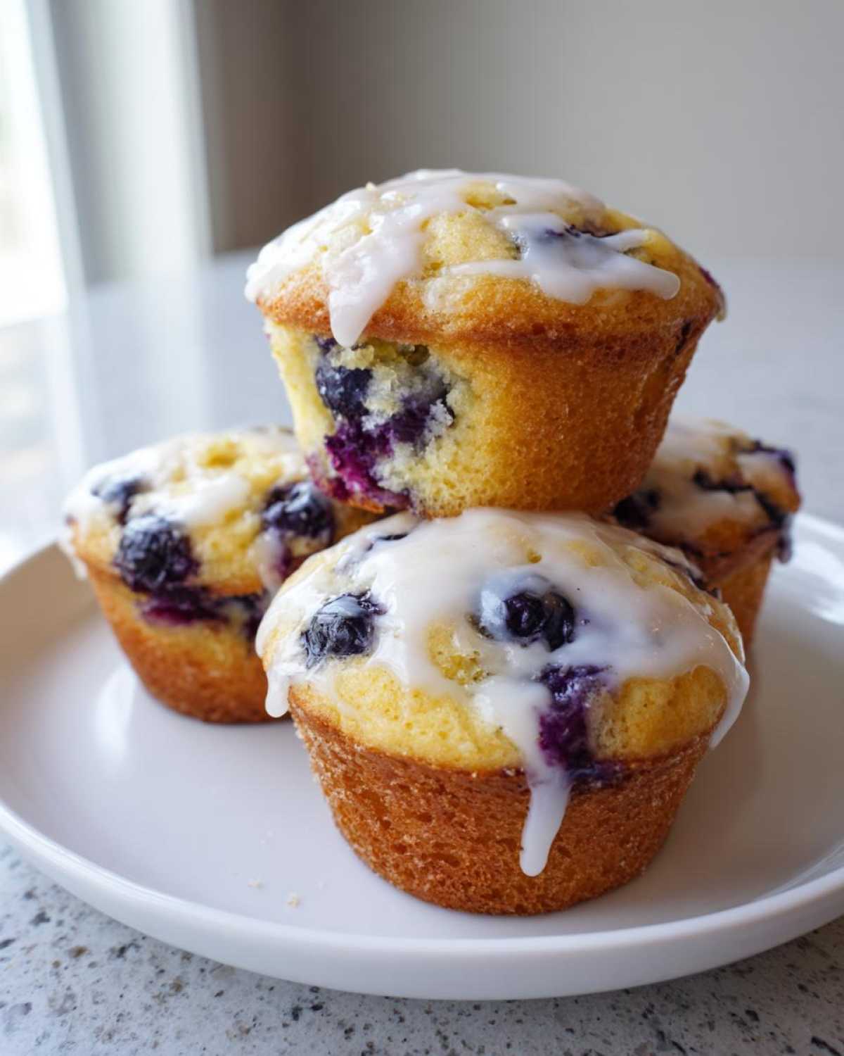 Close-up of four freshly baked Lemon Blueberry Muffins stacked on a white plate, topped with a sweet white glaze.
