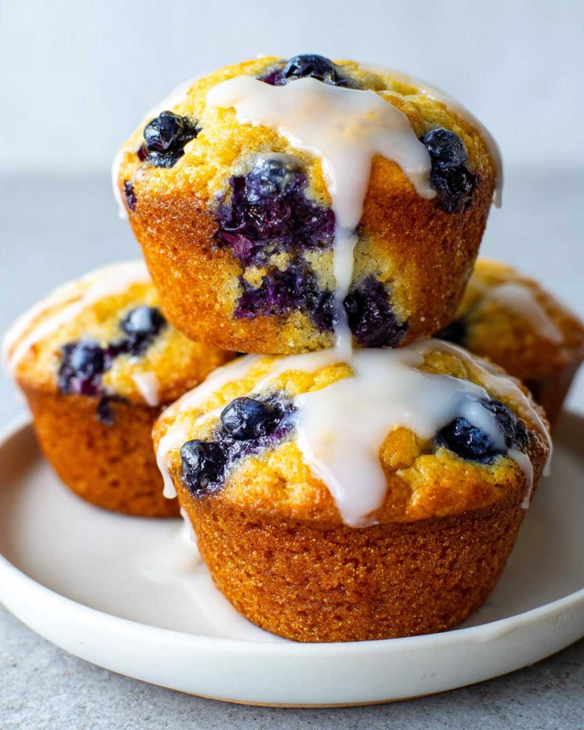 A stack of three golden Lemon Blueberry Muffins topped with a thick, white lemon glaze.