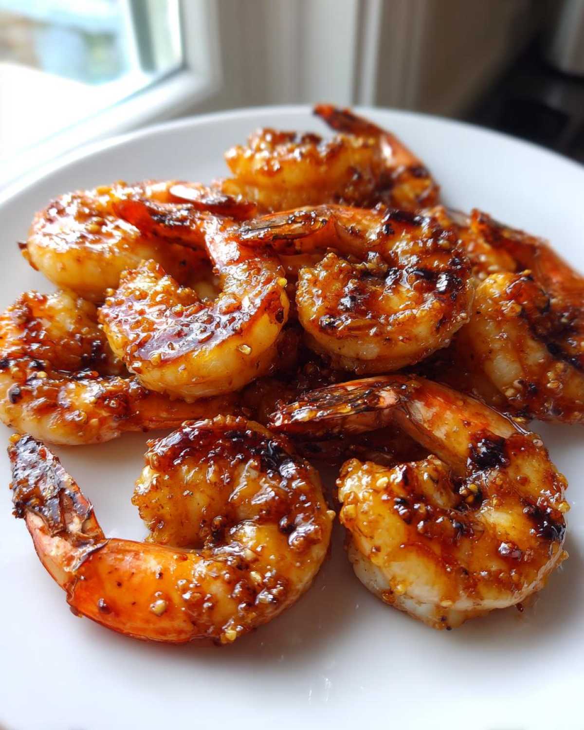 A close-up shot of several pieces of glossy, glazed Honey Garlic Grilled Shrimp piled on a white plate.