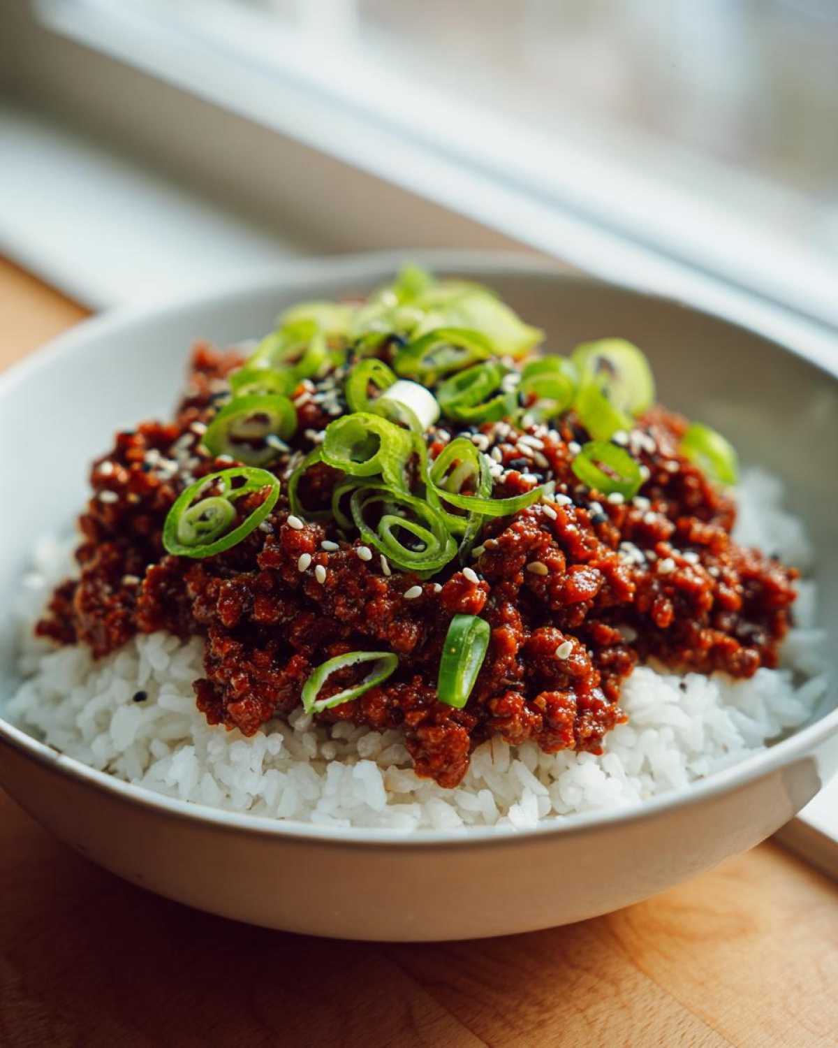 A close-up of a white bowl filled with white rice topped generously with spicy Korean Ground Beef Bowl and garnished with sliced green onions and sesame seeds.