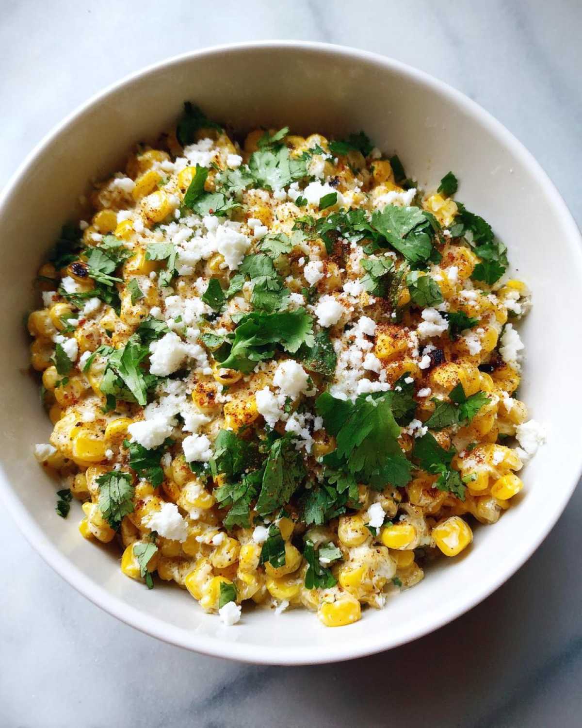 A close-up overhead shot of Mexican Street Corn Salad Esquites in a white bowl, topped with cotija cheese, cilantro, and chili powder.