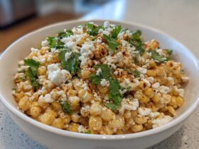A close-up of a white bowl filled with Mexican Street Corn Salad Esquites, topped with cotija cheese, cilantro, and chili powder.