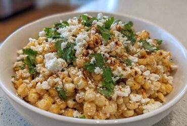 A close-up of a white bowl filled with Mexican Street Corn Salad Esquites, topped with cotija cheese, cilantro, and chili powder.
