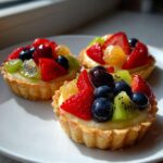 Three vibrant Mini Fruit Tarts topped with fresh strawberries, blueberries, kiwi, and glaze on a white plate.