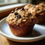 A close-up of a freshly baked Oatmeal Banana Chocolate Chip Muffin topped with oats and chocolate chips.