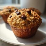 A close-up of a freshly baked Oatmeal Banana Chocolate Chip Muffin topped with oats and chocolate chips on a white plate.