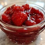 Close-up of whole strawberries suspended in thick, glossy syrup, representing Old-Fashioned Strawberry Preserves in a small glass jar.