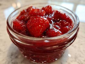 Close-up of whole strawberries suspended in thick, glossy syrup, representing Old-Fashioned Strawberry Preserves in a small glass jar.