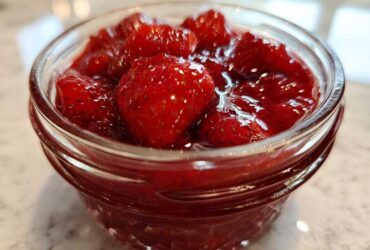 Close-up of whole strawberries suspended in thick, glossy syrup, representing Old-Fashioned Strawberry Preserves in a small glass jar.