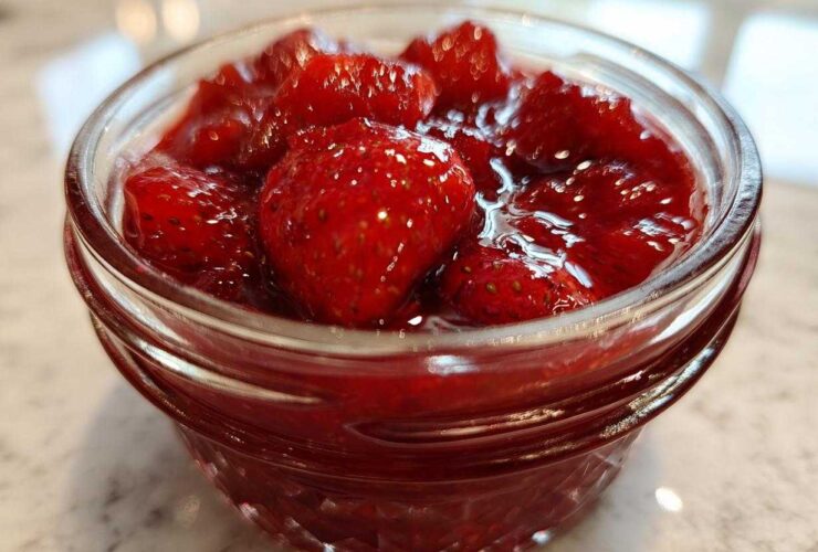 Close-up of whole strawberries suspended in thick, glossy syrup, representing Old-Fashioned Strawberry Preserves in a small glass jar.