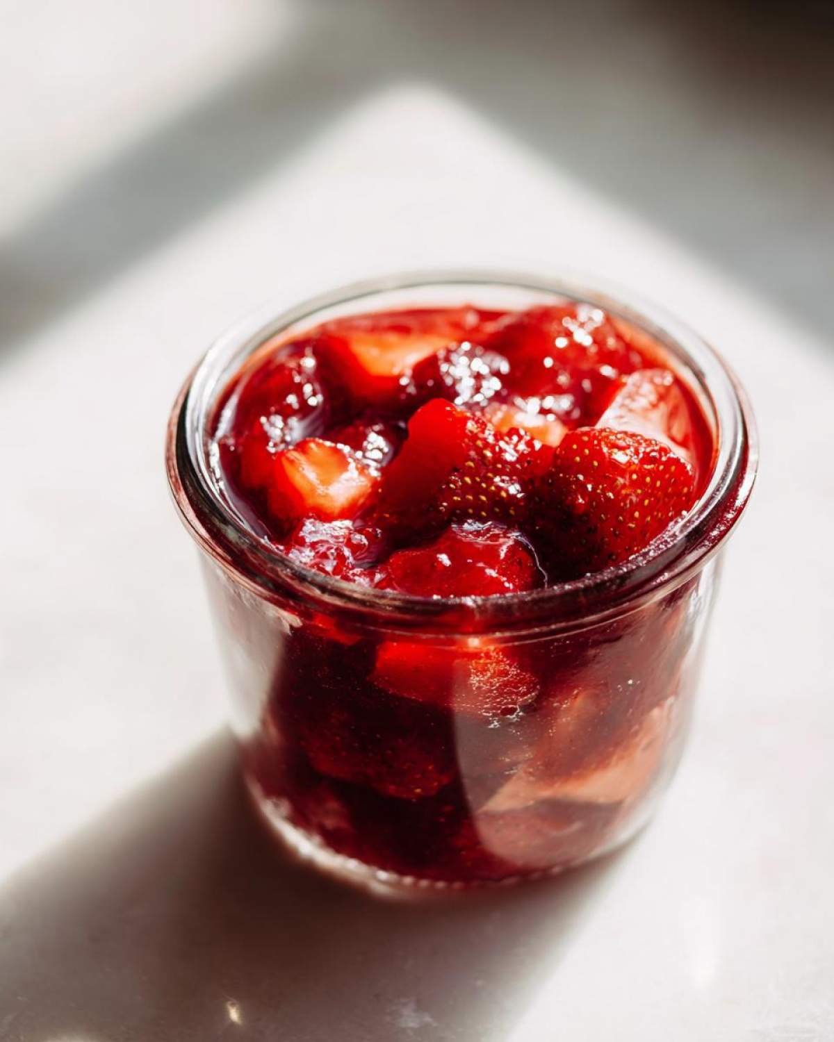 A glass jar filled with chunky Old-Fashioned Strawberry Preserves, glistening in bright sunlight.